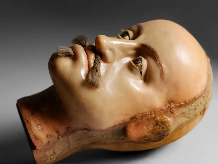 Eine realistische Kopfmaske mit einem blassen Teint, schmalen Lippen, und Augen, die leicht geöffnet sind. Sie hat einen gepflegten Schnurrbart und das Gesicht wirkt detailliert und lebensecht, aus einem hartem Material gefertigt. Der Hintergrund ist grau und neutral.