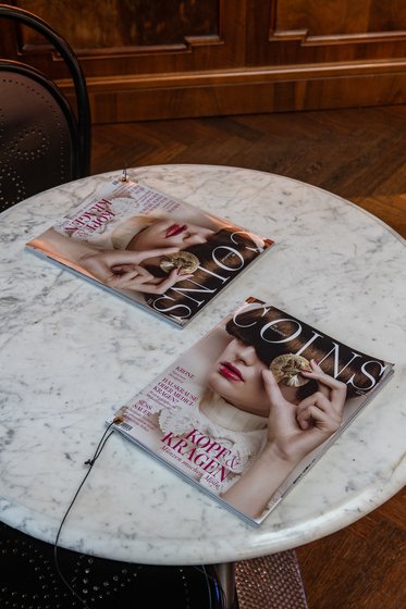Zwei Magazine liegen auf einem runden Marmortisch. Eines zeigt einen glamourösen Look mit einer Hand, die eine Münze hält, und das andere hat ein ähnliches Design. Der Tisch ist von Holz umgeben, was eine elegante Atmosphäre schafft.
