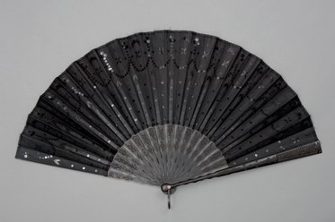 Ein schwarzer Fächer ist geöffnet, der aus mehreren schmalen Lamellen besteht. Seine Oberfläche ist mit feinen Verzierungen und glitzernden Punkten geschmückt. Der Fächer strahlt Eleganz und Raffinesse aus und ist auf einem grauen Hintergrund platziert.