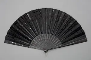 Ein schwarzer Fächer ist geöffnet, der aus mehreren schmalen Lamellen besteht. Seine Oberfläche ist mit feinen Verzierungen und glitzernden Punkten geschmückt. Der Fächer strahlt Eleganz und Raffinesse aus und ist auf einem grauen Hintergrund platziert.