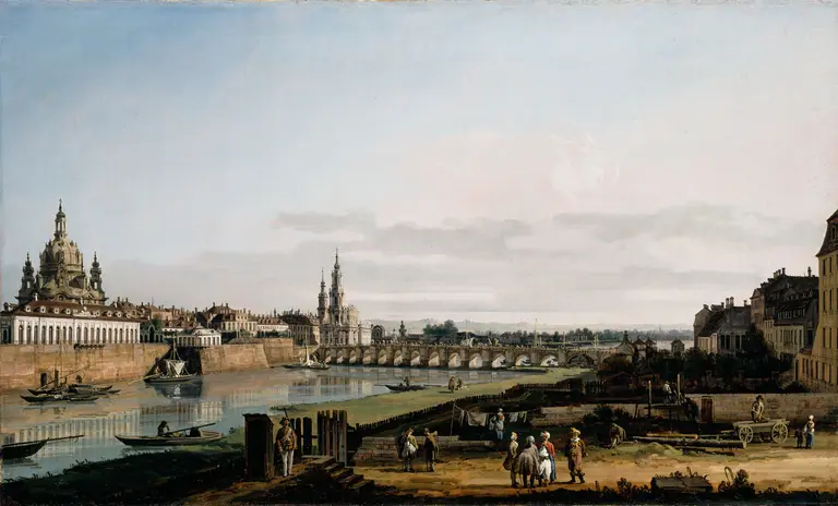 A panoramic view of a riverbank cityscape, showcasing architectural features like domes and spires. People are engaging in various activities along the river, with boats visible in the water. The scene is set against a clear blue sky, highlighting the city's serene environment.