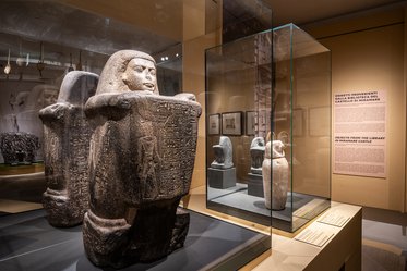 Die Abbildung zeigt eine Museumsansicht mit antiken Skulpturen in Vitrinen. Im Vordergrund steht eine große, steinerne Statue mit hieroglyphischen Inschriften. Im Hintergrund sind kleinere Reliefe und weitere Statuen zu sehen. Beleuchtung unterstreicht die Details der Objekte in der Ausstellung.