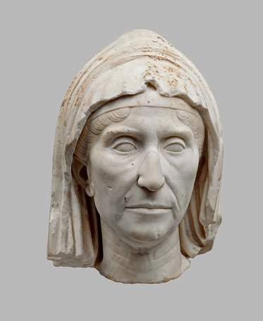 Eine antike Marmorbüste einer Frau mit einem einfachen Kleid und einem Kopftuch. Ihr Gesicht hat einen ernsten Ausdruck mit gut definierten Zügen. Die Skulptur weist Abnutzungserscheinungen auf, was auf ihr Alter hinweist. Der Hintergrund ist einfarbig grau.
