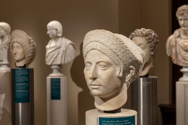 Eine Skulptur eines Kopfes mit geflochtenem Haar, umgeben von weiteren antiken Köpfen auf Sockeln. Die Figuren zeigen unterschiedliche Gesichtsausdrücke und Frisuren, und sind Teil einer Ausstellung in einem Museum. Der Hintergrund ist neutral gehalten.