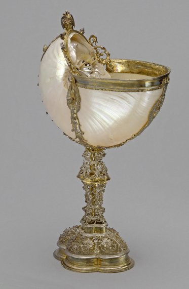 Ein kunstvoll gestalteter Becher aus Muschelschale und Silber. Der Becher hat eine runde Form und ist auf einem verzierten Fuß aus Silber montiert, der reich mit filigranen Details geschmückt ist. Die Muschel hat eine glänzende Oberfläche und wirkt elegant und geschmackvoll.