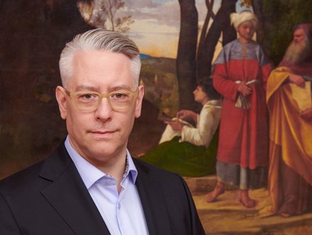 Ein Mann mit grauen Haaren und Brille trägt einen schwarzen Anzug und schaut direkt in die Kamera. Im Hintergrund sind historische Figuren in traditioneller Kleidung zu sehen, die in einer malerischen Landschaft stehen.