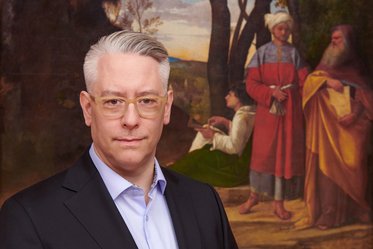 Ein Mann mit grauen Haaren und Brille trägt einen schwarzen Anzug und schaut direkt in die Kamera. Im Hintergrund sind historische Figuren in traditioneller Kleidung zu sehen, die in einer malerischen Landschaft stehen.