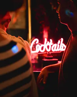 Ein neonschimmerndes Schild mit der Aufschrift „Cocktails“ leuchtet in einem dunklen Raum. Im Vordergrund sind Teile von Personen zu sehen, darunter eine Hand und ein gestreifter Ärmel. Die Atmosphäre wirkt einladend und gemütlich.
