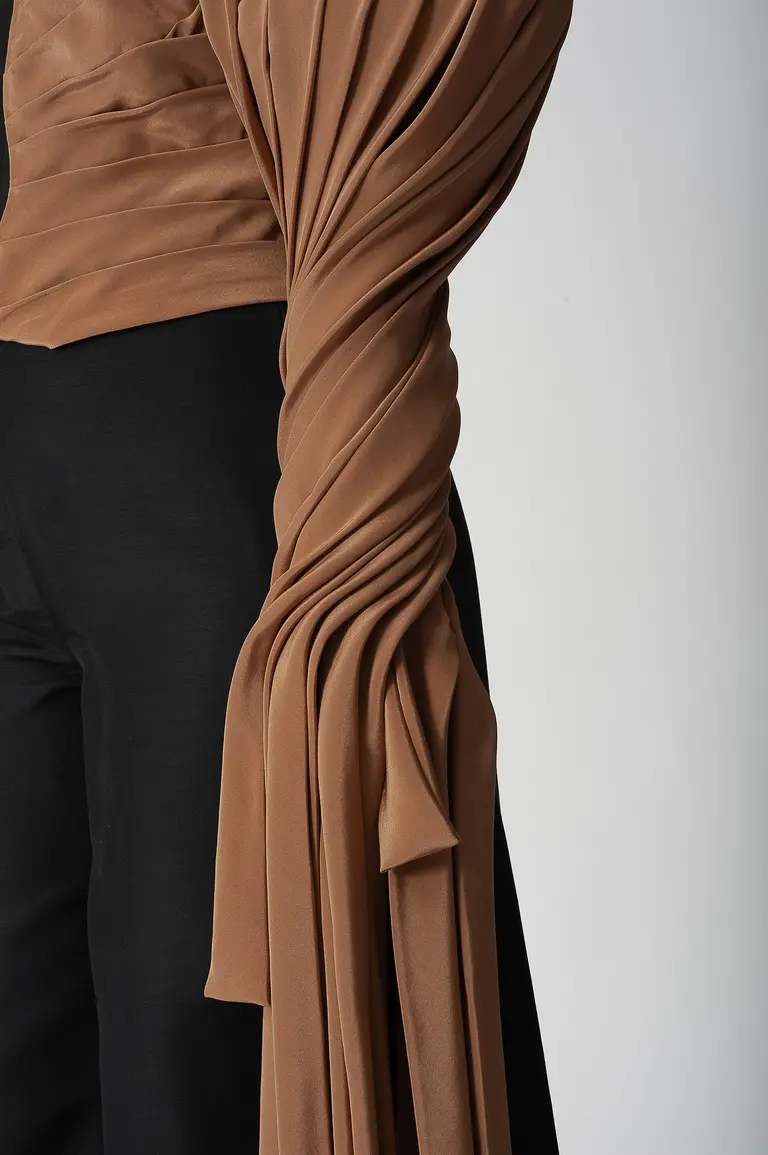 A close-up view of a person wearing a stylish outfit featuring a fitted, draped top in a warm brown color, with intricate fabric folds. The bottom part shows black pants, creating a contrast with the top's texture and color.