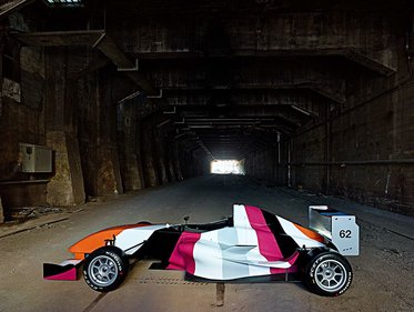 Ein auffälliges Rennauto mit einer bunten, abstrakten Lackierung steht in einem langen, dunklen Tunnel. Das Licht am Ende des Tunnels ist sichtbar, während die Wände aus Beton und Verfall bestehen. Die Nummer 62 ist an der Seite des Fahrzeugs angebracht.