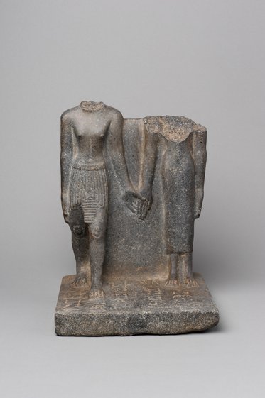Eine antike Steinfigur, die zwei gehauene Menschen darstellt, die Händchen haltend nebeneinander stehen. Die Köpfe und Teile des Körpers sind beschädigt und fehlen. Die Figur zeigt typische Merkmale ägyptischer Kunst, wie die detaillierte Darstellung der Kleidung.