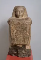 Eine alte ägyptische Statue aus Stein, die eine Menschengestalt darstellt. Die Figur hat einen schmalen Kopf und trägt eine kunstvolle Inschrift auf der Vorderseite. Die Statue steht auf einer roten Podestfläche und zeigt detaillierte Reliefs auf der Oberfläche.
