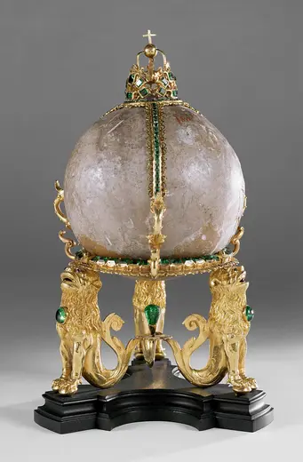 Eine verschnörkelte goldene Globuskugel auf einer schwarzem Sockel, flankiert von zwei goldenen Löwenfiguren. Die Kugel ist mit grünen Edelsteinen und einer Krone geschmückt. Der Hintergrund ist schlicht und hebt das kunstvolle Objekt hervor.