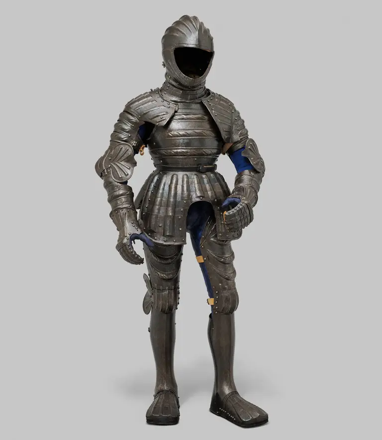 Alterstext: Eine metallene Rüstung steht aufrecht. Sie besteht aus mehreren beweglichen Teilen und hat einen vollständig geschlossenen Helm ohne Gesichtsausschnitt. Die Rüstung wirkt detailreich und zeigt eine Kombination aus Schutz und Ästhetik. Der Hintergrund ist grau.