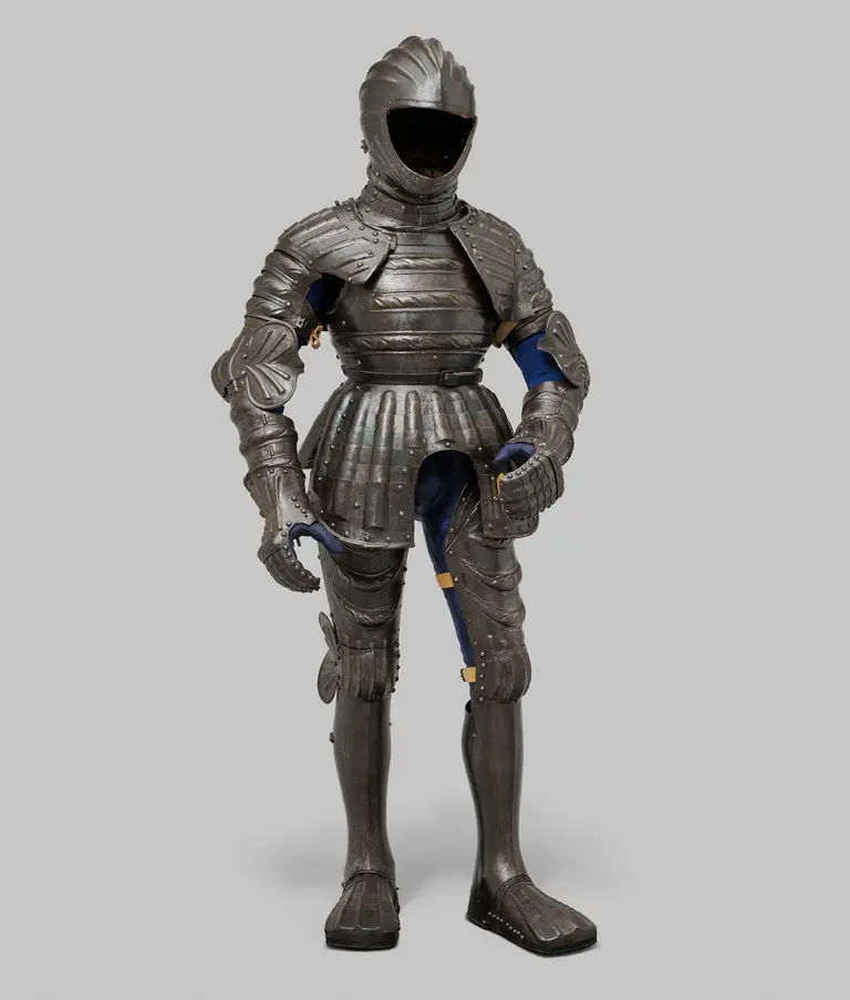 A full suit of medieval armor stands upright against a gray background. The armor features detailed plates covering the torso, arms, and legs, with a helmet that has a smooth, closed faceplate. The suit is metallic gray and shows signs of craftsmanship typical of historical armor designs.
