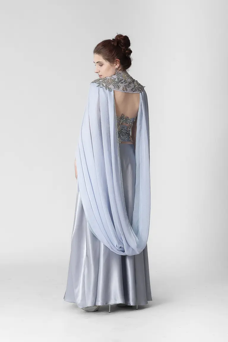 A woman is standing with her back to the viewer, wearing a floor-length, silver gown with a flowing light blue cape. The outfit features intricate embellishments on the upper back and shoulders, complementing the elegance of the dress. The background is plain and neutral.