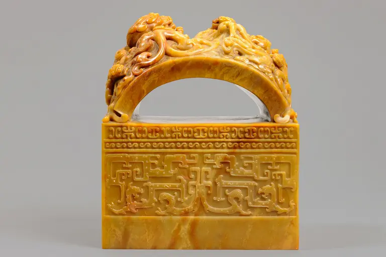 Das Bild zeigt einen kunstvoll geschnitzten jadeartigen Stempel mit einem geschwungenen Griff, der mit Drachenmotiven verziert ist. Die Basis des Stempels hat eine ornierte Oberfläche. Der Stempel ist in warmen Gelbtönen gehalten und vor einem grauen Hintergrund platziert.