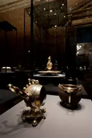 Im Vordergrund befinden sich zwei kunstvoll gestaltete silberne Gefäße auf einem dunklen Untergrund. Links steht eine Kanne mit dekorativem Henkel, rechts ein tiefes Gefäß mit einem runden Rand. Im Hintergrund sind weitere Objekte in einer Vitrine zu erkennen.