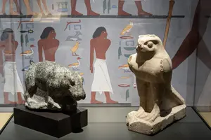 Zwei antike Statuen sind auf einer Glasfläche präsentiert. Links steht eine schwarze Steinfigur eines Tieres, rechts eine hellere Statue eines Vogels mit menschlichen Zügen, beide vor einer Wand mit ägyptischen Hieroglyphen und Figuren.