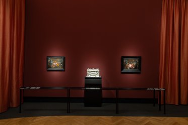 Eine Galeriewand mit einem dunklen roten Hintergrund. Zwei gemalte Bilder in schwarzen Rahmen hängen an der Wand. In der Mitte steht ein Podest mit einem dekorativen Objekt. Vor der Wand sind lange Tische platziert, die Informationen präsentieren. Die Beleuchtung ist sanft und einladend.