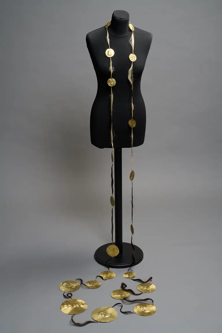 A black mannequin displays a long, decorative gold necklace featuring circular pendants. Additional circular pendants lie on the ground, enhancing the focus on the jewelry's intricate design. The background is a neutral gray, emphasizing the jewelry's details.