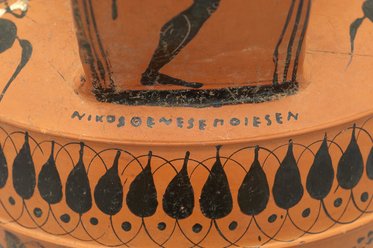 Die Abbildung zeigt eine antike griechische Vase mit schwarzer Bemalung. Darauf sind menschliche Figuren und dekorative Motive in Form von Tropfen und Kreisen zu sehen. Am Rand ist eine Inschrift, die den Namen „Nikosorenes” zeigt.