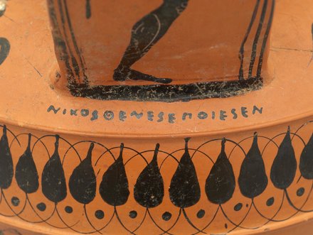Die Abbildung zeigt eine antike griechische Vase mit schwarzer Bemalung. Darauf sind menschliche Figuren und dekorative Motive in Form von Tropfen und Kreisen zu sehen. Am Rand ist eine Inschrift, die den Namen „Nikosorenes” zeigt.