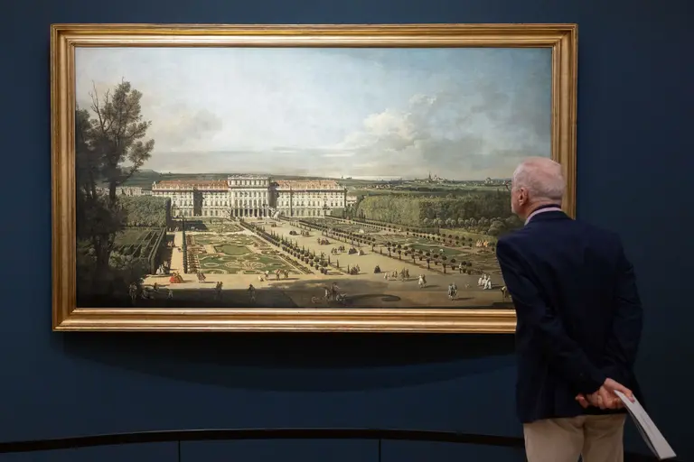 A person stands in front of a large painting depicting an expansive formal garden with manicured hedges and pathways, alongside a grand building. The scene captures a sunny day with a blue sky and scattered clouds, set within a gallery.