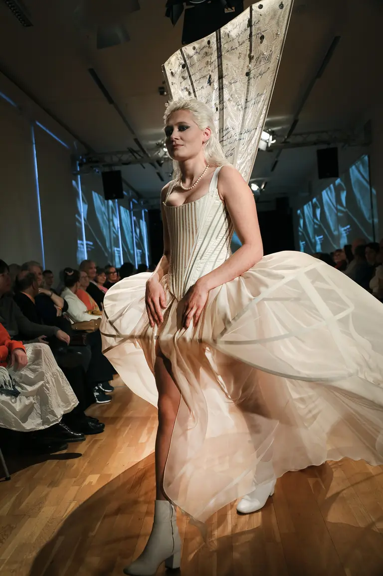 A model walks the runway in a striking fashion piece featuring a corset top and a voluminous, layered skirt. The outfit includes an elaborate design element resembling a fan or wing structure. The audience is visible in the background, watching the show.
