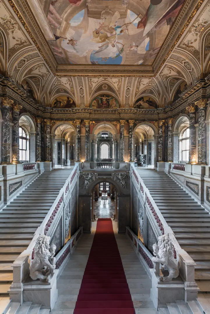 Ein prächtiger Treppenaufgang in einem historischen Gebäude. Die breite Treppe mit rotem Teppich wird von kunstvollen Säulen und Statuen flankiert. Oberhalb befindet sich eine reich verzierte Decke mit Malereien. Licht strömt durch große Fenster, die den Raum erhellen.