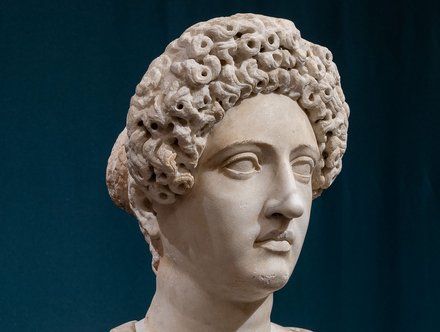 Eine antike Marmorbüste mit einem detailreich gestalteten Kopf, der lockige Haare hat. Der Gesichtsausdruck ist ruhig und konzentriert, mit betonten Augen und einer klaren Nase. Der Hintergrund ist dunkel, was die Figur hervorhebt.