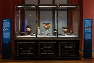 Vitrine in einem Museum mit antiken Vasen und Schalen. Die Objekte sind auf einem weißen Tisch präsentiert, umgeben von Glas. Links und rechts stehen Informationssäulen mit Beschreibungen. Der Hintergrund ist dunkelrot, was die Ausstellungsstücke hervorhebt.