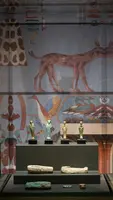 Eine Vitrine mit vier kleinen antiken Statuen in unterschiedlichen Farben auf einem schwarzen Sockel. Im Hintergrund ist eine Wandmalerei mit verschiedenen Tiermotiven, darunter ein Hund, und hieroglyphischen Zeichen zu sehen.