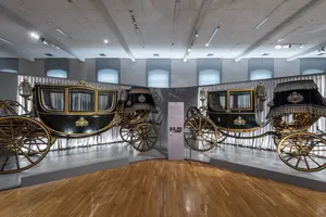 Das Bild zeigt zwei prunkvolle Kutschen in einem Ausstellungsraum. Die Kutschen sind schwarz lackiert und mit goldenen Verzierungen geschmückt. Sie stehen nebeneinander auf einem Holzfußboden, umgeben von sanftem Licht und Vorhängen. Eine Informationssäule befindet sich zwischen ihnen.