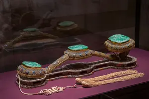 Ein prächtiges ornamental dekoriertes Objekt mit drei grünen Jadeplatten, eingefasst in ein kunstvolles Muster aus Gold und blauen Edelsteinen. Es liegt auf einer samtigen Oberfläche, neben am Boden befestigten Quasten, die die Eleganz des Artefakts unterstreichen.