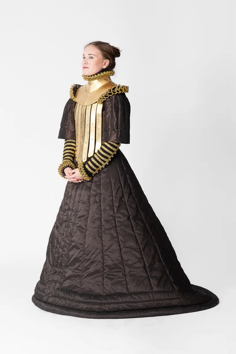A woman stands in profile, wearing a distinctive black gown with a voluminous skirt and a high, ornate collar. The dress features gold accents and structured sleeves. The background is plain white, emphasizing the unique design of the outfit.