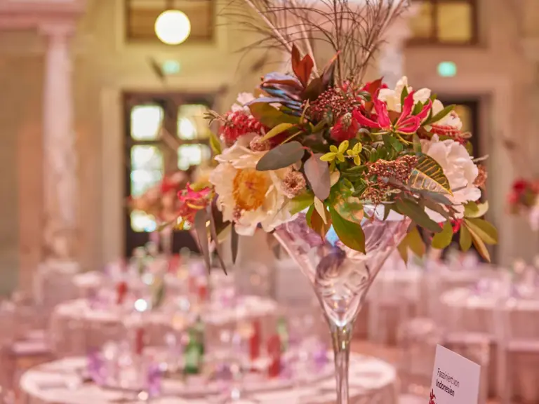 A tall, elegant floral arrangement featuring colorful flowers and peacock feathers sits in a crystal vase at the center of a beautifully set dining table, surrounded by chair backs. The backdrop hints at an ornate venue, creating an atmosphere suitable for a sophisticated event.