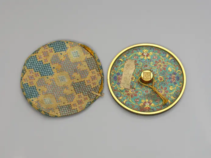 Ein runder Handspiegel in einem prächtigen, blauen und goldfarbenen Etui. Der Spiegel hat eine aufwendige, florale Dekoration und einen goldenen Griff. Das Etui ist mit einem bunten, geometrischen Muster versehen. Der Spiegel ist geöffnet und zeigt seine kunstvolle Gestaltung.