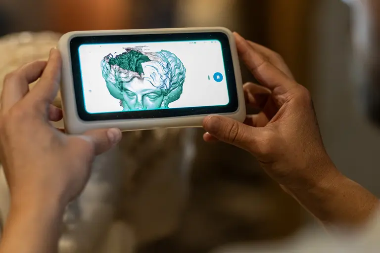 Eine Person hält ein mobiles Gerät, das eine digitale Darstellung eines menschlichen Kopfes und Gehirns zeigt. Die Grafik ist in verschiedenen Grüntönen gehalten und zeigt Details der Gehirnstruktur. Der Hintergrund zeigt eine Skulptur, die unscharf ist und den Fokus auf das Gerät lenkt.