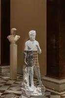 Eine moderne Skulptur zeigt eine teilweise transparente Figur eines Mannes mit einem Netz an ihrem Bauch. Im Hintergrund steht eine weiße Büste eines antiken Mannes auf einem Sockel. Der Boden ist mit klassischen Fliesenmustern gestaltet.