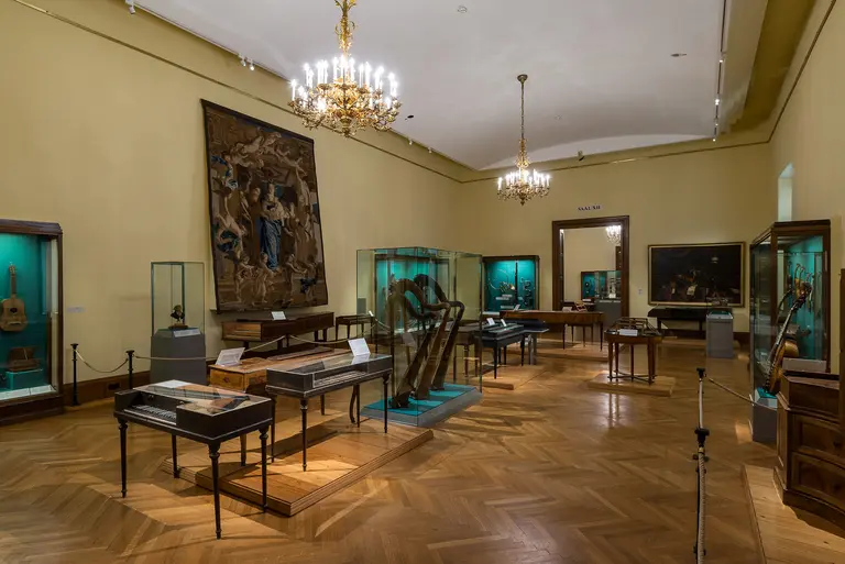 Das Bild zeigt einen Ausstellungsraum in einem Museum mit verschiedenen Musikinstrumenten in Vitrinen. Im Hintergrund hängen Gemälde und ein großer Wandteppich. Die Wände sind gelb gestrichen, und eine Deckenleuchte sorgt für Beleuchtung. Der Boden ist aus Holz.