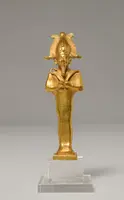 Eine goldene Figur eines Pharaos mit einem kunstvollen Kopfschmuck und in traditioneller Kleidung. Sie hält in beiden Händen Symbole der Macht. Die Statue steht auf einem transparenten Sockel vor einem neutralen Hintergrund.