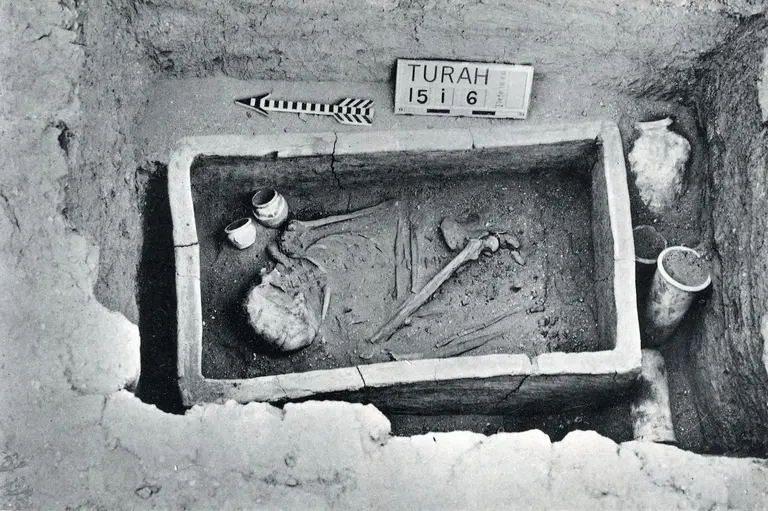 Schwarz-Weiß-Foto eines Grabes aus der Ausgrabungsstätte in Turah, 1910. Zu sehen ist ein rechteckiger Grabschacht mit Skelettresten und beigegebenen Keramikgefäßen. Ein Schild mit der Aufschrift 