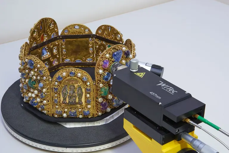 Goldene Krone mit bunten Edelsteinen auf einem schwarzen Drehteller, ein technisches Gerät mit der Aufschrift WITEC 457nm und einem Warnschild