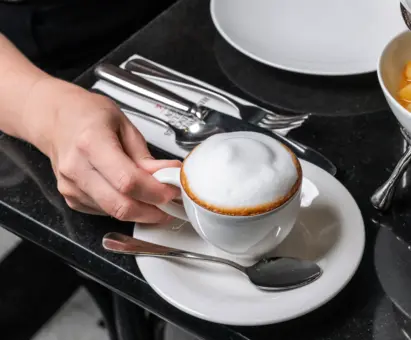 Eine Hand hält eine kleine Tasse mit schaumigem Kaffee, die auf einem weißen Unterteller steht. Daneben liegt ein kleiner Löffel. Der Hintergrund zeigt einen eleganten Tisch mit Besteck und Geschirr.