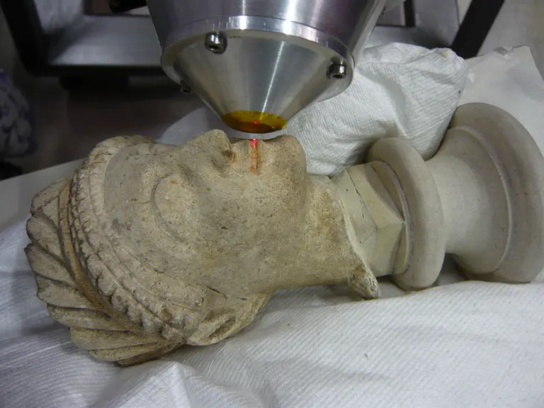 Ein Lasergerät ist auf das Gesicht einer beschädigten Skulptur gerichtet, die auf einer weißen Unterlage liegt. Der Laserstrahl ist sichtbar und trifft auf die Nase der Skulptur, während sie vorsichtig behandelt wird.