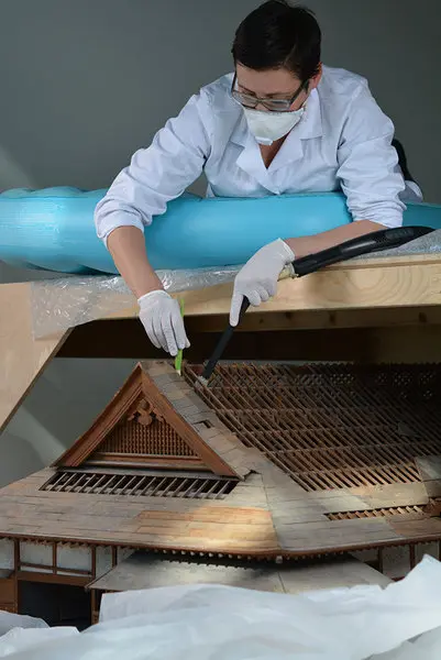 Eine Person in Schutzkleidung arbeitet an einem detaillierten Modell eines Gebäudedachs. Sie verwendet Werkzeuge, um vorsichtig an der Struktur zu arbeiten. Der Arbeitsplatz ist ordentlich und zeigt ein handwerkliches Restaurierungsprojekt.