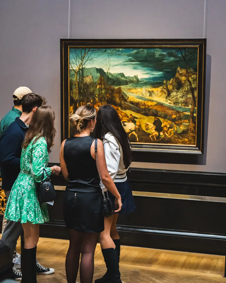 Eine Gruppe von fünf Personen steht in einem Museum vor einem Gemälde. Die Menschen betrachten das Kunstwerk, das eine Landschaft zeigt, während sie sich unterhalten. Die Farbpalette des Gemäldes umfasst Grüntöne und Erdtöne.