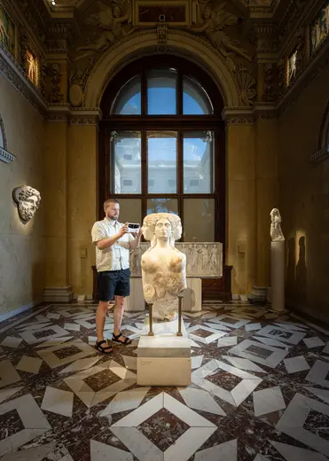 Ein Besucher steht vor einer antiken Marmorbüste in einem Museum. Der Raum hat hohe Fenster und eine dekorative Fußbodenmusterung. Der Mann hält ein Smartphone, während er die Statue betrachtet. Im Hintergrund sind weitere Skulpturen und Reliefformen sichtbar.