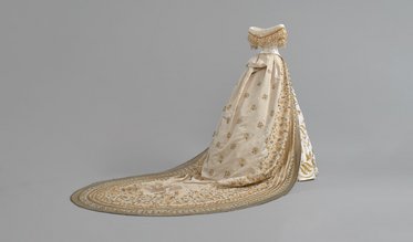Ein opulent gestaltetes, bodenlanges Kleid in hellem Beige mit kunstvollen Goldstickereien. Der Rock hat eine breite Bordüre, die sich elegant um die untere Kante wölbt. Das Oberteil ist mit einer dekorativen Halslinie und einer wiegenden Schleife verziert.